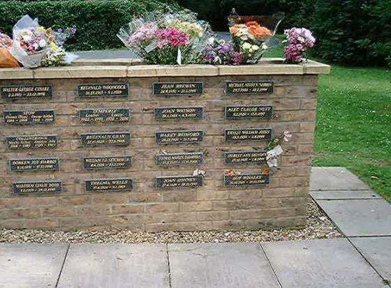 Waist height brick wall displaying 16 granite plaques in 4x4 layout with bouquets of flowers placed in the top of the wall